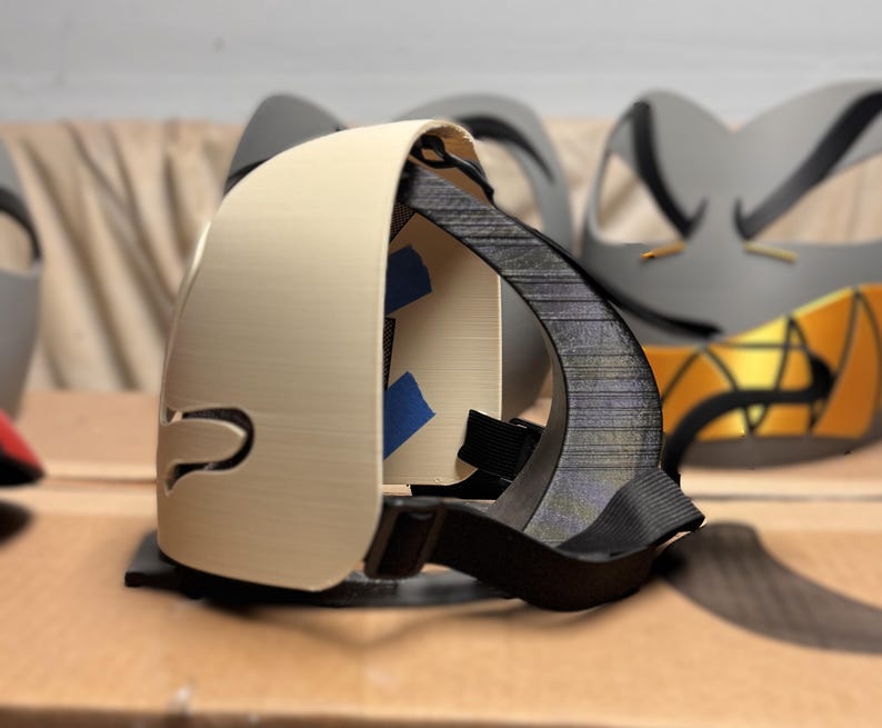 May include: A beige 3D printed mask with a smooth exterior and a black interior frame. The mask features a black adjustable elastic strap attached to the sides and small pieces of blue painter's tape visible on the inner structure of the mask.