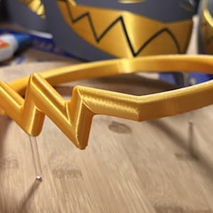 May include: A gold-colored headband featuring a prominent, angular zigzag design at the front. The headband has a smooth, rounded band structure and a distinct, sharp geometric shape centered on the top, crafted with a visible 3D printed texture.