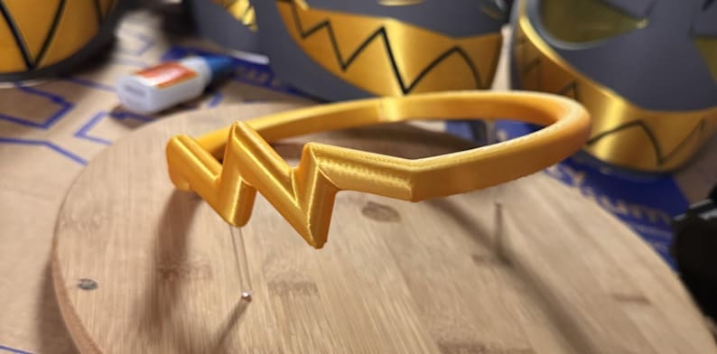 May include: A gold-colored headband featuring a prominent, angular zigzag design at the front. The headband has a smooth, rounded band structure and a distinct, sharp geometric shape centered on the top, crafted with a visible 3D printed texture.