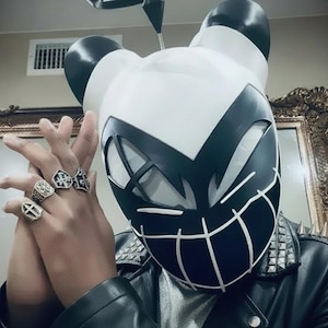 Puede incluir: Una máscara en blanco y negro con un diseño de rostro estilizado. La máscara tiene un patrón en forma de V y una boca sonriente. La persona lleva una chaqueta de cuero negra con tachuelas plateadas y varios anillos.