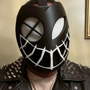 May include: Black mask with white accents, featuring a stitched grin and eye details. The mask has a strap and is worn with a black leather jacket with silver spikes on the shoulders. The mask's design is reminiscent of a comic book character.