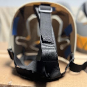 May include: The interior of a protective mask featuring a beige curved shell with white structural padding. Black adjustable nylon straps are attached to the frame with small blue rectangular adhesive tabs securing the internal components in place.