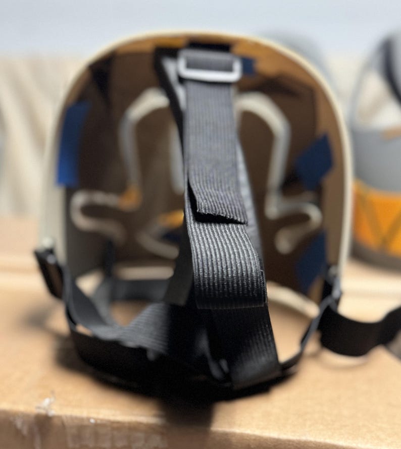 May include: The interior of a protective mask featuring a beige curved shell with white structural padding. Black adjustable nylon straps are attached to the frame with small blue rectangular adhesive tabs securing the internal components in place.