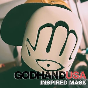 May include: A white mask featuring a black hand and eye graphic, worn under a mustard yellow baseball cap. The text GODHANDUSA INSPIRED MASK is printed in black and red at the bottom of the image, clearly visible against the dark background.
