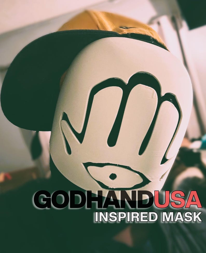 May include: A white mask featuring a black hand and eye graphic, worn under a mustard yellow baseball cap. The text GODHANDUSA INSPIRED MASK is printed in black and red at the bottom of the image, clearly visible against the dark background.