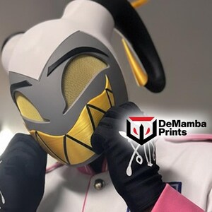 May include: A white, gray, and yellow character mask featuring a gold halo, mesh eye coverings, and a wide, jagged yellow grin. The mask is worn with black gloves featuring white graphic details and a white jacket with pink trim and silver buttons.