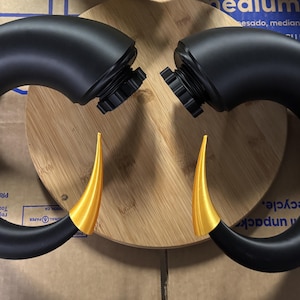 May include: A pair of curved, matte black horns with pointed, metallic gold tips. Each horn features a circular, threaded black base attachment point at the top, designed for secure mounting. The horns are positioned symmetrically, curving inward toward each other.