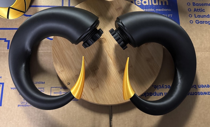 May include: A pair of curved, matte black horns with pointed, metallic gold tips. Each horn features a circular, threaded black base attachment point at the top, designed for secure mounting. The horns are positioned symmetrically, curving inward toward each other.