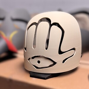 May include: A cream-coloured, dome-shaped mask featuring a cutout design of a hand with an eye symbol at the base. The mask has a black mesh backing visible through the cutouts and is mounted on a small black base, showcasing a textured surface.