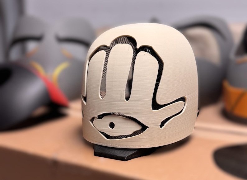 May include: A cream-coloured, dome-shaped mask featuring a cutout design of a hand with an eye symbol at the base. The mask has a black mesh backing visible through the cutouts and is mounted on a small black base, showcasing a textured surface.