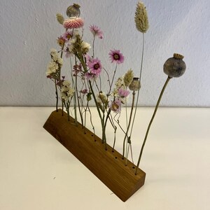 Op de afbeelding: Een houten bloemenstandaard met 10 gaten voor gedroogde bloemen. De standaard is gemaakt van lichtbruin hout en heeft een gebogen vorm. De bloemen zijn een mix van roze, wit en bruin.