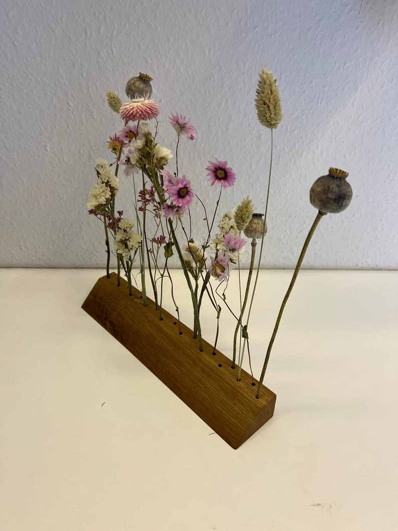 Op de afbeelding: Een houten bloemenstandaard met 10 gaten voor gedroogde bloemen. De standaard is gemaakt van lichtbruin hout en heeft een gebogen vorm. De bloemen zijn een mix van roze, wit en bruin.