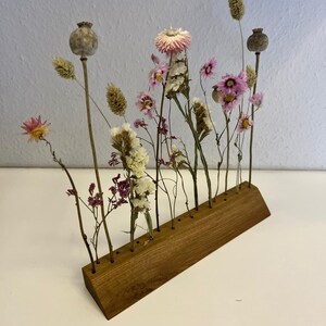 Op de afbeelding: Een houten bloemenstandaard met gedroogde bloemen in verschillende tinten roze en wit. De standaard heeft meerdere gaten om de stelen van de bloemen vast te houden.
