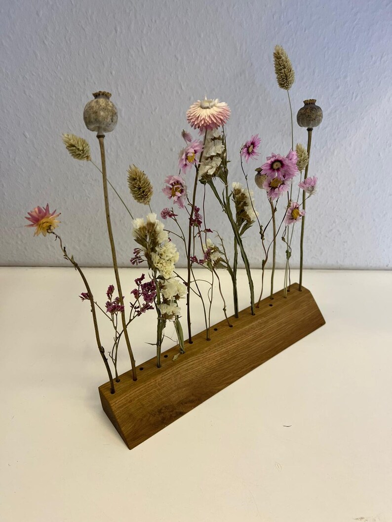 Op de afbeelding: Een houten bloemenstandaard met gedroogde bloemen in verschillende tinten roze en wit. De standaard heeft meerdere gaten om de stelen van de bloemen vast te houden.