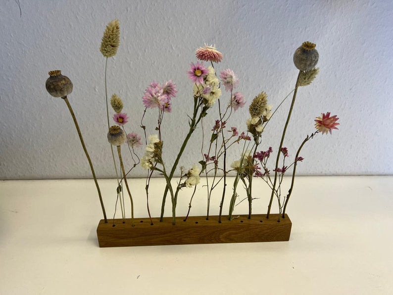 Op de afbeelding: Gedroogde bloemenarrangement in een houten standaard met 12 sleuven. De bloemen zijn een mix van roze, wit en bruin. De standaard is gemaakt van natuurlijk hout.