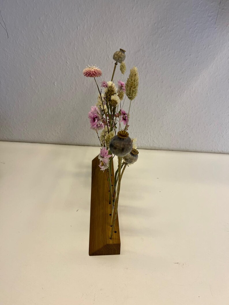 Op de afbeelding: Een houten bloemenhouder met een driehoekige basis en meerdere gaten voor gedroogde bloemen. De houder is gevuld met gedroogde bloemen in tinten roze, wit en bruin.