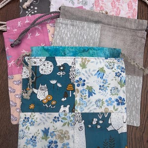 May include: A collection of rectangular fabric drawstring bags featuring various patchwork designs. The bags display patterns including floral motifs, woodland creatures like owls and bears, and abstract botanical prints in shades of blue, pink, grey, and white with braided cord closures.