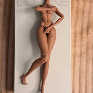 Pode incluir: Um corpo de boneca articulado de cor bronzeada com um torso sem cabeça, braços cruzados e uma perna dobrada. A boneca está sobre uma superfície retangular branca com a palavra "DOARE" impressa.