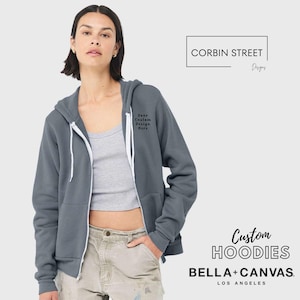 May include: A slate gray zip-up hoodie with a white zipper and drawstring. The hoodie has a small embroidered design on the chest that reads "Your Custom Design Here". The words "Custom Hoodies" and "BELLA + CANVAS" are also visible.