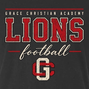 May include: A dark gray t-shirt featuring the text GRACE CHRISTIAN ACADEMY in white, followed by the word LIONS in large, distressed red block letters. Below this, the word football is written in white script, with a red and white G logo.