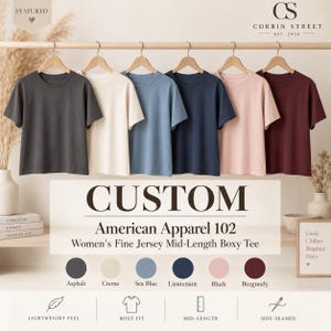 May include: A collection of six short-sleeved t-shirts hanging on a wooden rack in asphalt, creme, sea blue, lieutenant, blush, and burgundy colors. The text reads CUSTOM American Apparel 102 Fine Jersey Mid-Length Boxy Tee with color labels below.