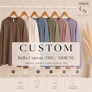 May include: A collection of six long-sleeve crew neck t-shirts hanging on wooden hangers in colors including vintage brown, soft cream, steel blue, heather military green, heather mauve, and heather dark lavender, labeled Bella Canvas 3501.