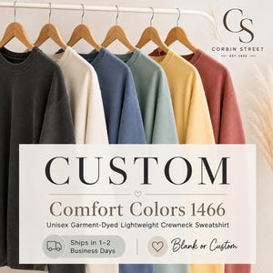 May include: A row of six crewneck sweatshirts hanging on wooden hangers in various colors including black, cream, blue, sage green, yellow, and terracotta. The text reads CUSTOM Comfort Colors 1466 Unisex Garment-Dyed Lightweight Crewneck Sweatshirt.
