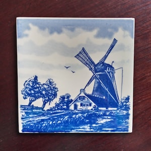 May include: Blue and white ceramic tile featuring a depiction of a Dutch windmill, a small house, and trees. The tile has a blue and white background with a cloudy sky.