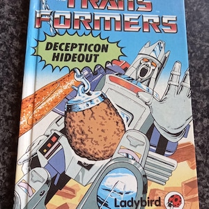 May include: A hardback book titled The Transformers Decepticon Hideout with a blue cover featuring a large illustration of a robot character, a brown textured object, and the Ladybird logo with a small red ladybug icon in the bottom corner.