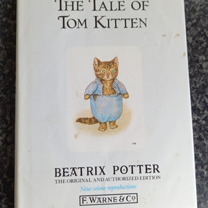 Puede incluir: La portada del libro "La Historia de Tom Kitten" con un gatito marrón y blanco que lleva una camisa azul y está de pie en un cuenco azul. La portada del libro es blanca con texto negro que dice "La Historia de Tom Kitten" y "Beatrix Potter".