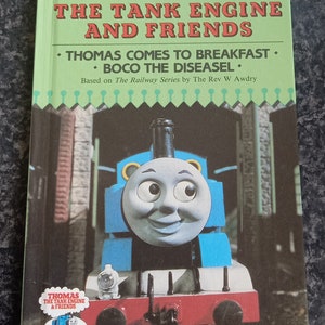 May include: A blue and red toy train engine with a smiling face. The engine is labeled "Thomas the Tank Engine & Friends". The book cover features the text "Thomas the Tank Engine and Friends" and the titles of two stories: "Thomas Comes to Breakfast" and "Boco the Diesease".