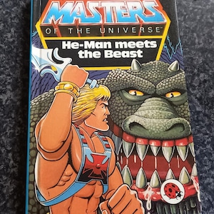 Może przedstawiać: Okładka starej książki Masters of the Universe przedstawiająca He-Mana, muskularnego mężczyznę o blond włosach, stojącego naprzeciwko zielonego, zębatego potwora. Tytuł książki brzmi "He-Man spotyka Bestię".