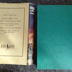 May include: An open vintage book with a cream-colored page featuring a list of titles from the "Biggles" series. The spine of a book with a colorful illustration is visible, along with a teal-colored book cover.