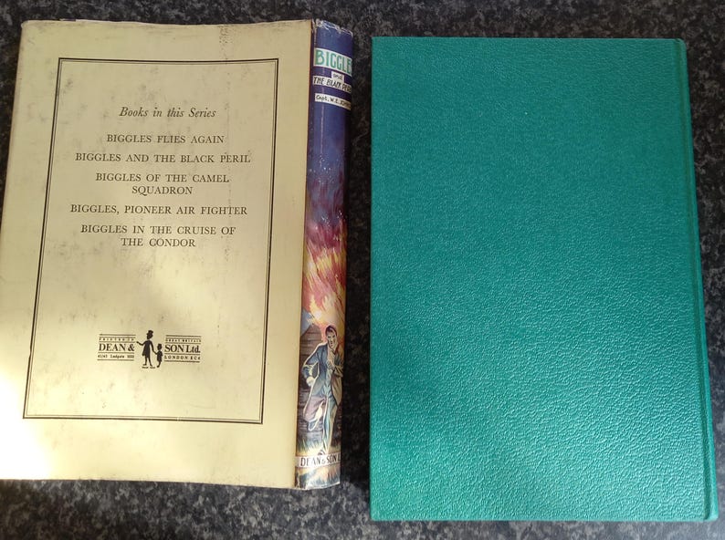 May include: An open vintage book with a cream-colored page featuring a list of titles from the "Biggles" series. The spine of a book with a colorful illustration is visible, along with a teal-colored book cover.