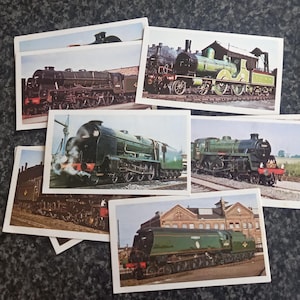 May include: A collection of vintage train cards featuring various steam locomotives. The cards showcase detailed images of green, black, and brown trains, capturing the essence of classic railway engineering. The cards are arranged on a dark, speckled surface.