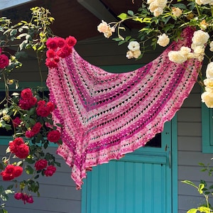 May include: A handmade, open-weave shawl in shades of pink and white hangs in front of a turquoise door. The shawl is draped between red and white roses, creating a vibrant contrast. The shawl's design features a geometric pattern.