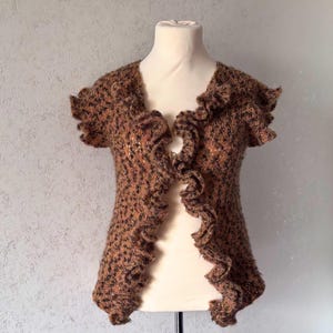 Könnte beinhalten: Eine braun-schwarze Strickweste mit Rüschenbesatz. Die Weste wird auf einer Schaufensterpuppe präsentiert und hat einen V-Ausschnitt. Der Strickstoff hat ein strukturiertes Aussehen, und die Rüschen verleihen dem Kleidungsstück ein dekoratives Element.