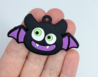 May include: A black bat-shaped charm with purple wings and a loop for hanging. The bat has large green eyes, white teeth, and a smiling purple mouth. This Halloween-themed charm is a fun accessory.