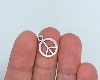 May include: A small, silver-coloured peace sign charm. The charm features the iconic peace symbol within a circular frame, with a small loop at the top for attaching to a chain or other jewellery. The charm is held between two fingers.