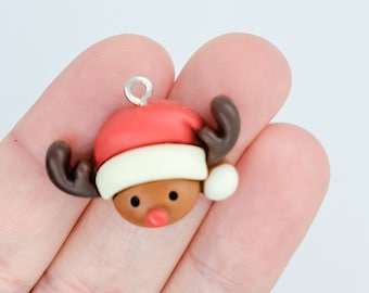 May include: A festive Christmas charm featuring a reindeer head wearing a red Santa hat with a white brim. The reindeer has brown antlers, a brown face, and a red nose. A silver loop is attached for hanging.