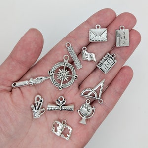 May include: A collection of silver charm pendants with school and education themes. The charms include a compass, a ruler, a book, a globe, a graduation cap, a pencil, a protractor, and a notebook.