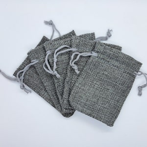 May include: A set of six small grey hessian drawstring bags. The bags are made of a woven fabric and have a light grey drawstring.