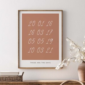 Könnte beinhalten: Ein gerahmter Druck mit terracottafarbenem Hintergrund und weißem Text. Der Text lautet "20 01 16", "16 03 17", "05 05 19", "10 03 21" und "THESE ARE THE DAYS".