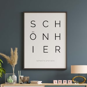 Könnte beinhalten: Ein schwarzer gerahmter Druck mit dem Wort "SCHÖNHIER" in weißer Schrift und den Koordinaten "50°58'3"N 6°57'25"E" in kleinerer weißer Schrift. Der Druck hängt an einer dunkelblauen Wand über einem Holzregal mit einer Vase, einer Holzskulptur und einem Stapel Büchern. Auf dem Regal befinden sich auch rosa Buchstabenblöcke, die "LOVE" buchstabieren.