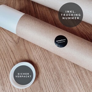 May include: A brown cardboard tube with a white end cap. The tube has a sticker that says "MyHappyPlace.co Let us tell your story". The tube also has a sticker that says "INKL. TRACKING NUMMER". A white lid with a grey circle that says "SICHER VERPACKT" is in the foreground.