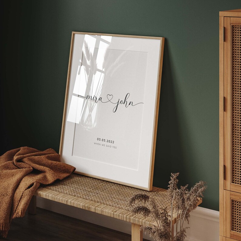 May include: A white framed print with the text "Mira & John" in cursive script with a heart symbol between the names. The date "03.03.2022" is printed below the names with the text "When we said yes" underneath the date.