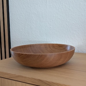 Könnte beinhalten: Eine flache, runde Holzschale in natürlichem Hellbraun mit sichtbarer Holzmaserung. Die Schale hat eine glatte, geschwungene Innen- und Außenfläche, die die organische Textur und die warmen Farbtöne des Holzmaterials zur Geltung bringt.