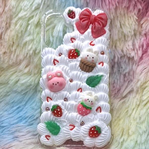 Könnte beinhalten: Transparente Handyhülle mit weißem Zuckerguss, roten Erdbeeren und niedlichen Tierfiguren verziert. Oben befindet sich eine rote Schleife. Der Text "Pingpan Studio" ist unten in Schreibschrift geschrieben. Die Hülle befindet sich auf einem bunten, flauschigen Hintergrund.