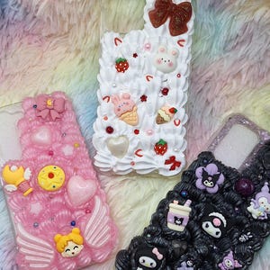 Op de afbeelding: Drie verschillende telefoonhoesjes versierd met kleurrijke 3D-versieringen. De eerste hoes is roze met een hart, een ster en een gevleugelde toverstaf. De tweede hoes is wit met een roze konijn, aardbeien en ijshoorns. De derde hoes is zwart met een zwarte kat, een paarse bloem en een zwart-witte mok.