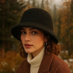 May include: A woman wearing a black cloche hat, a white turtleneck sweater, and a brown coat. The hat has a rounded crown and a downward-sloping brim. The coat is a warm, earthy brown color.
